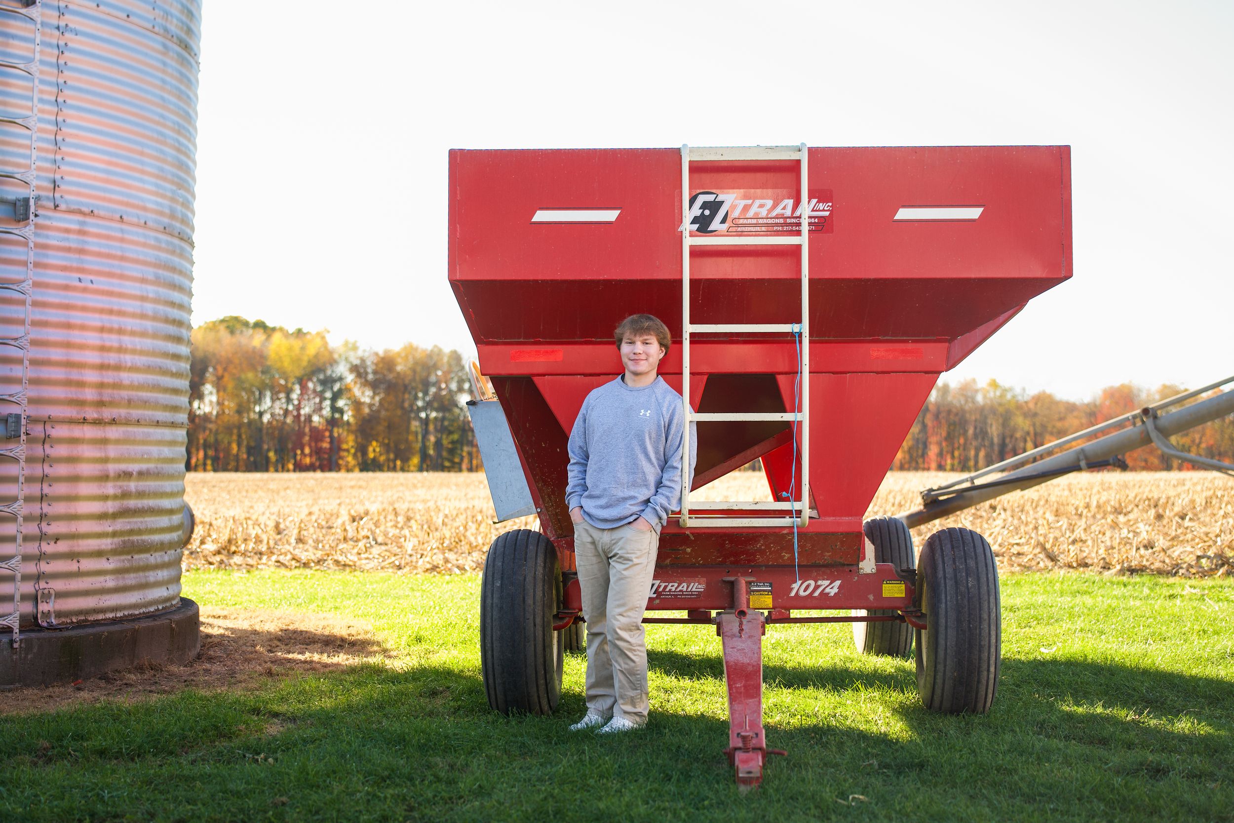 Person stands beside a large red farm grain wagon in a harvested corn field with metal silos visible nearby.