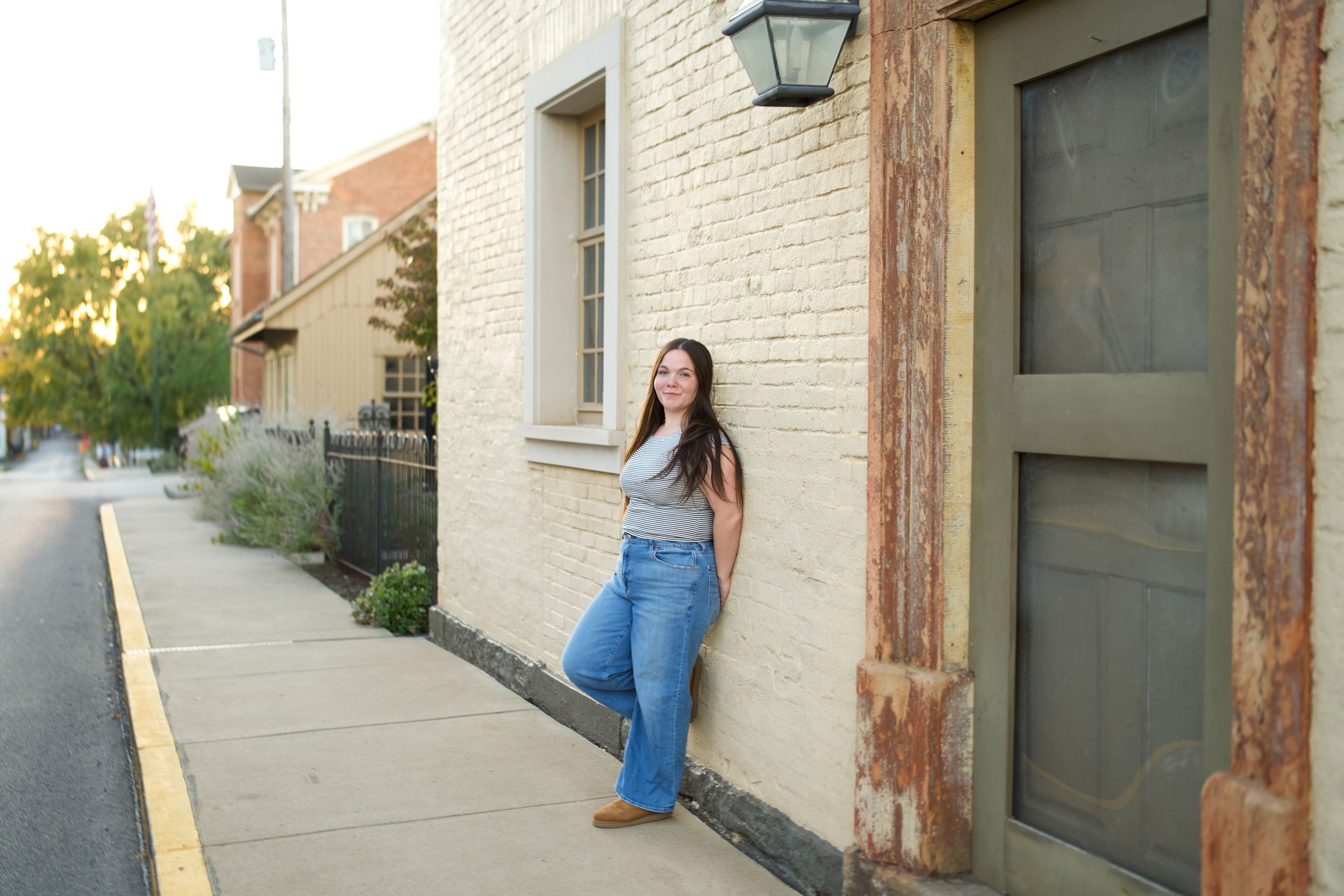 Young woman in jeans leans against a brick building on a sunny sidewalk in a small town street scene.
