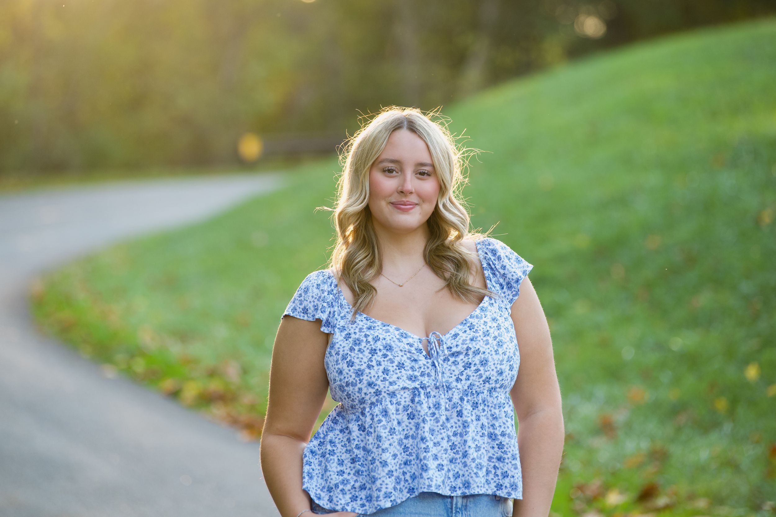 Woman wearing a blue floral top and jeans stands on a curving path surrounded by green grass in golden sunlight.