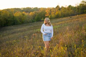 A person in a white sweater poses in a golden autumn meadow during sunset with rolling hills in the background.