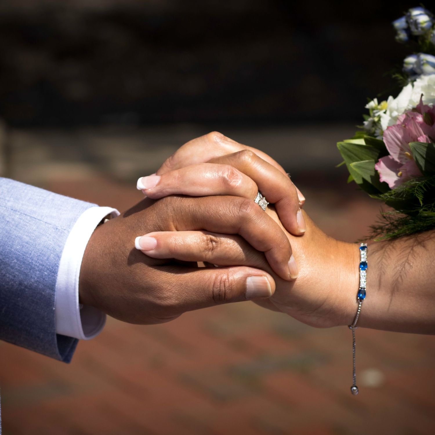 The couple holding hands with wedding rings and a delicate bracelet visible in a close-up shot.