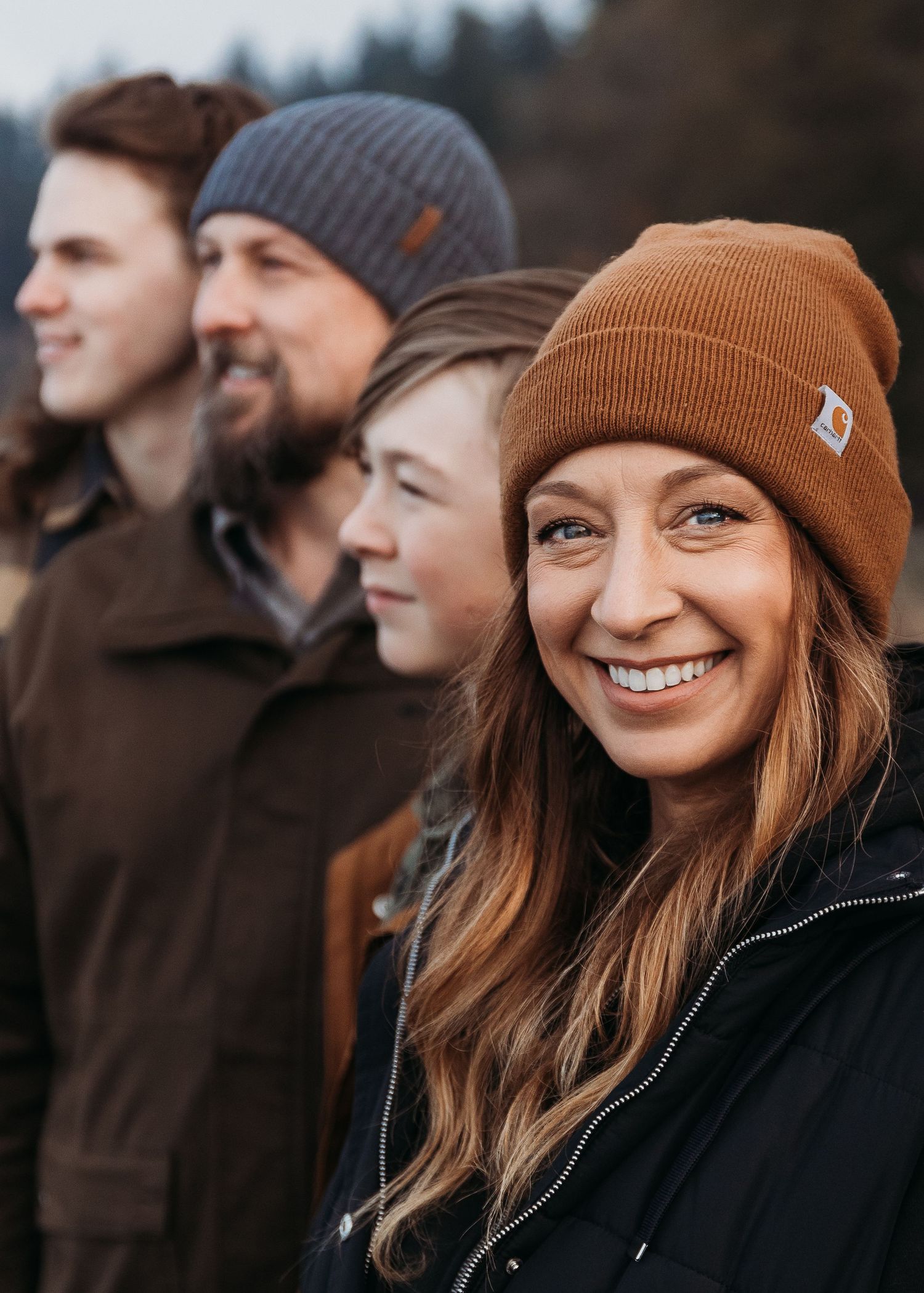 A group wearing winter beanies poses together outdoors with evergreen trees in the background.