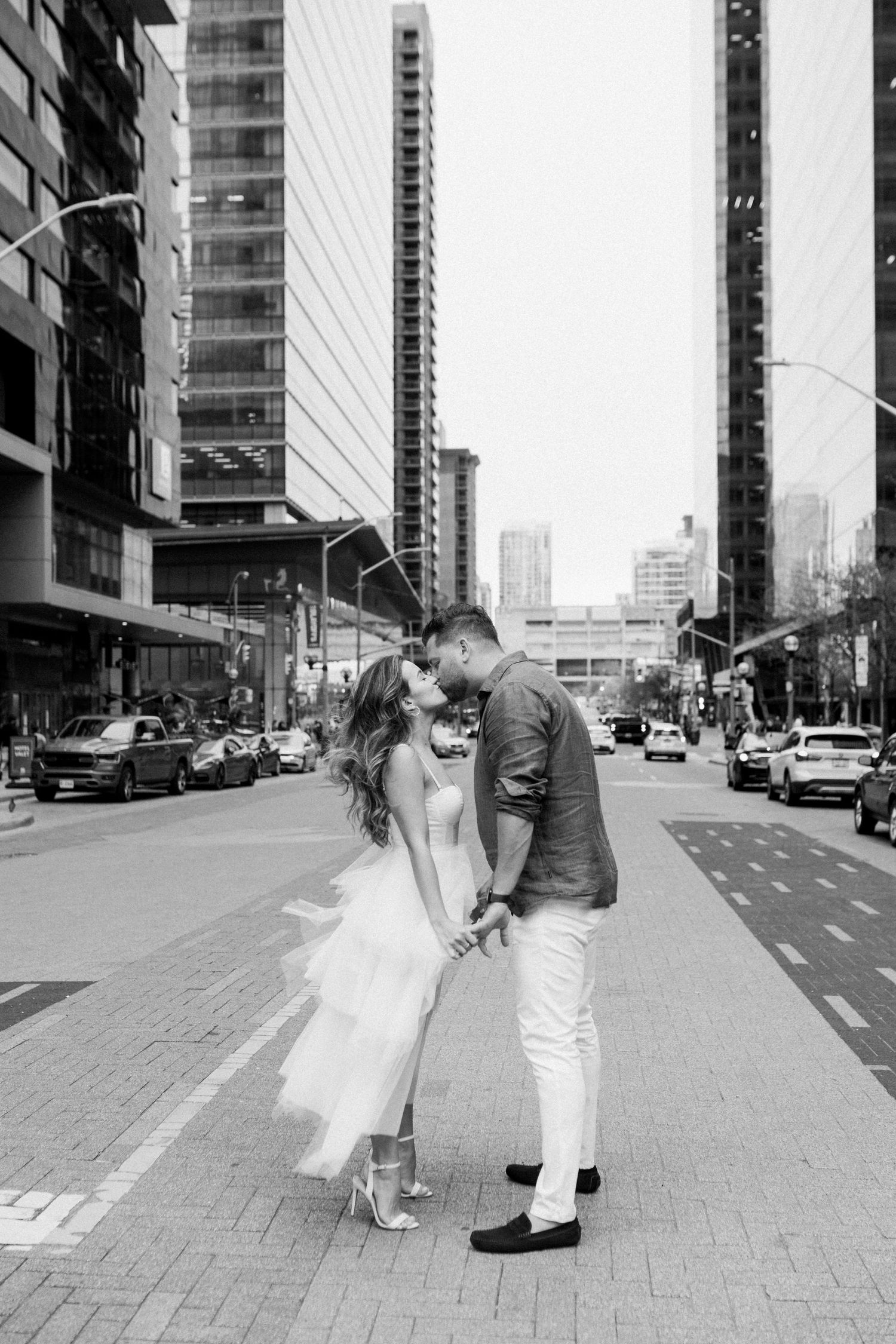 Romantic couple kissing on urban street with city buildings in black and white photography.