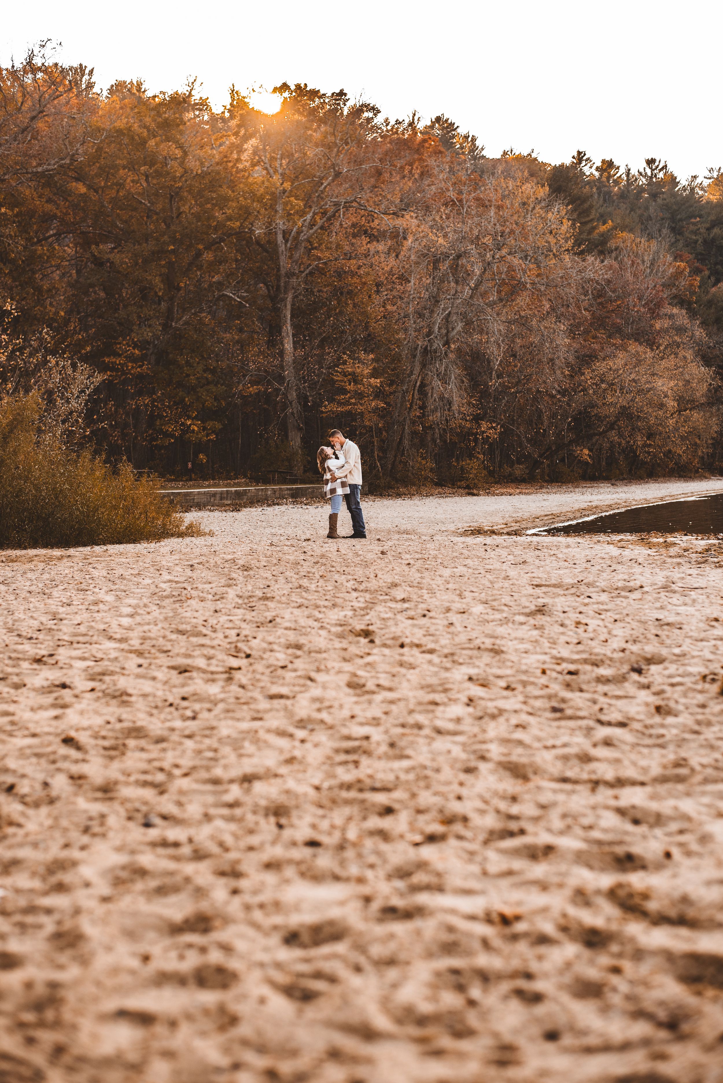 A couple embraces on a sandy beach path at sunset with autumn trees creating a warm golden backdrop.