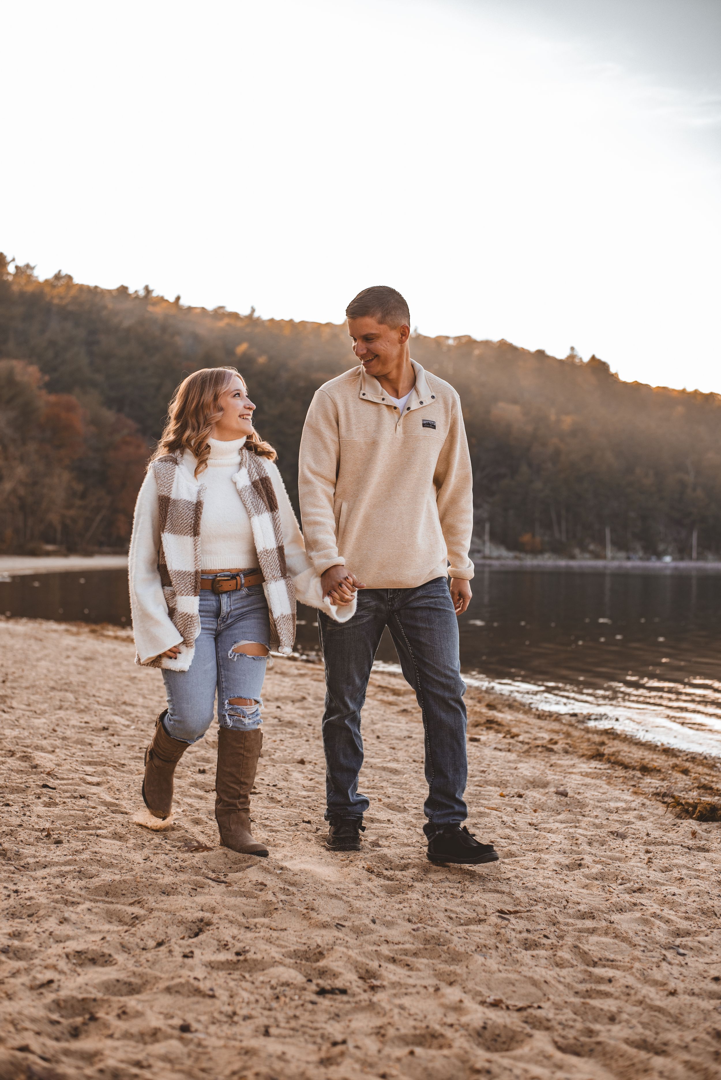 A couple walks hand in hand along a lakeside beach trail surrounded by fall foliage.