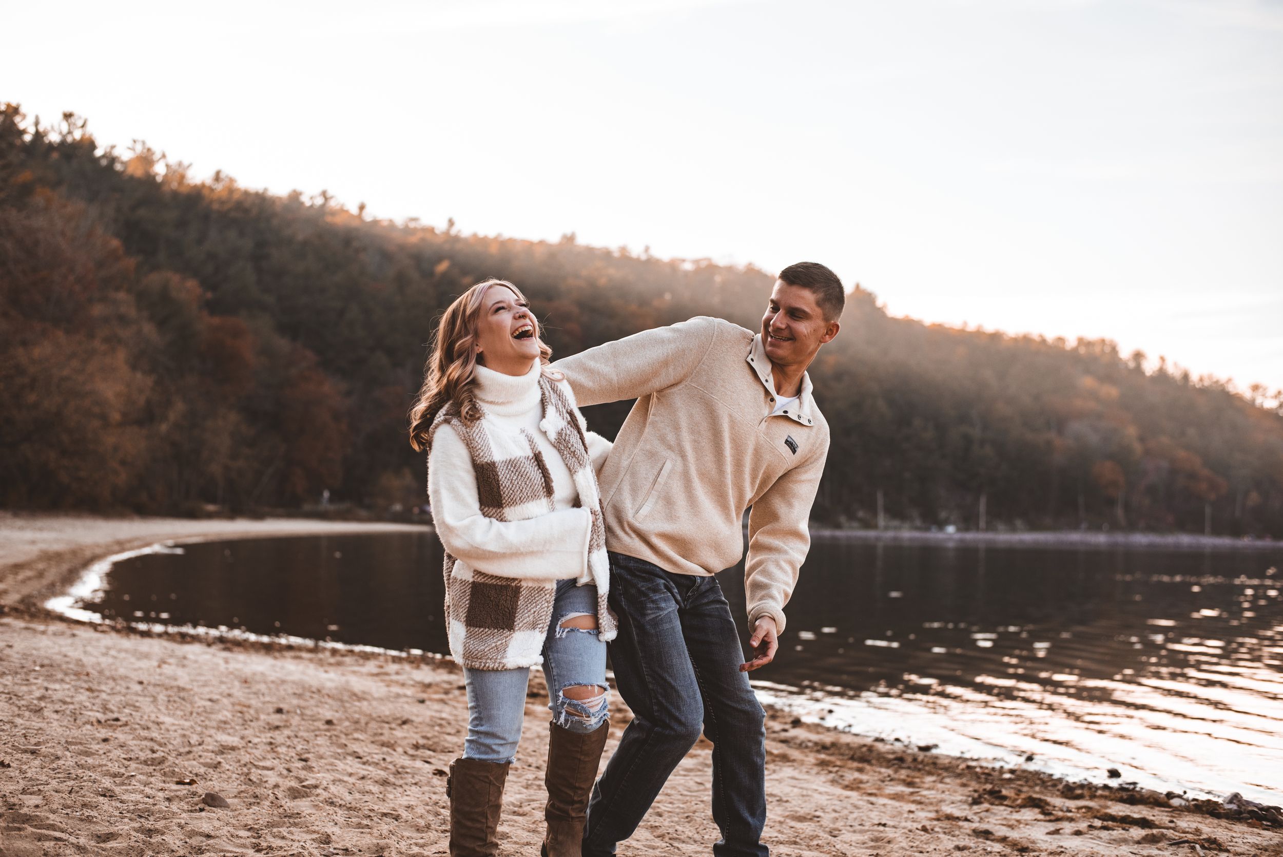 Playful couple laughing and dancing on a beach with autumn trees and lake in background.