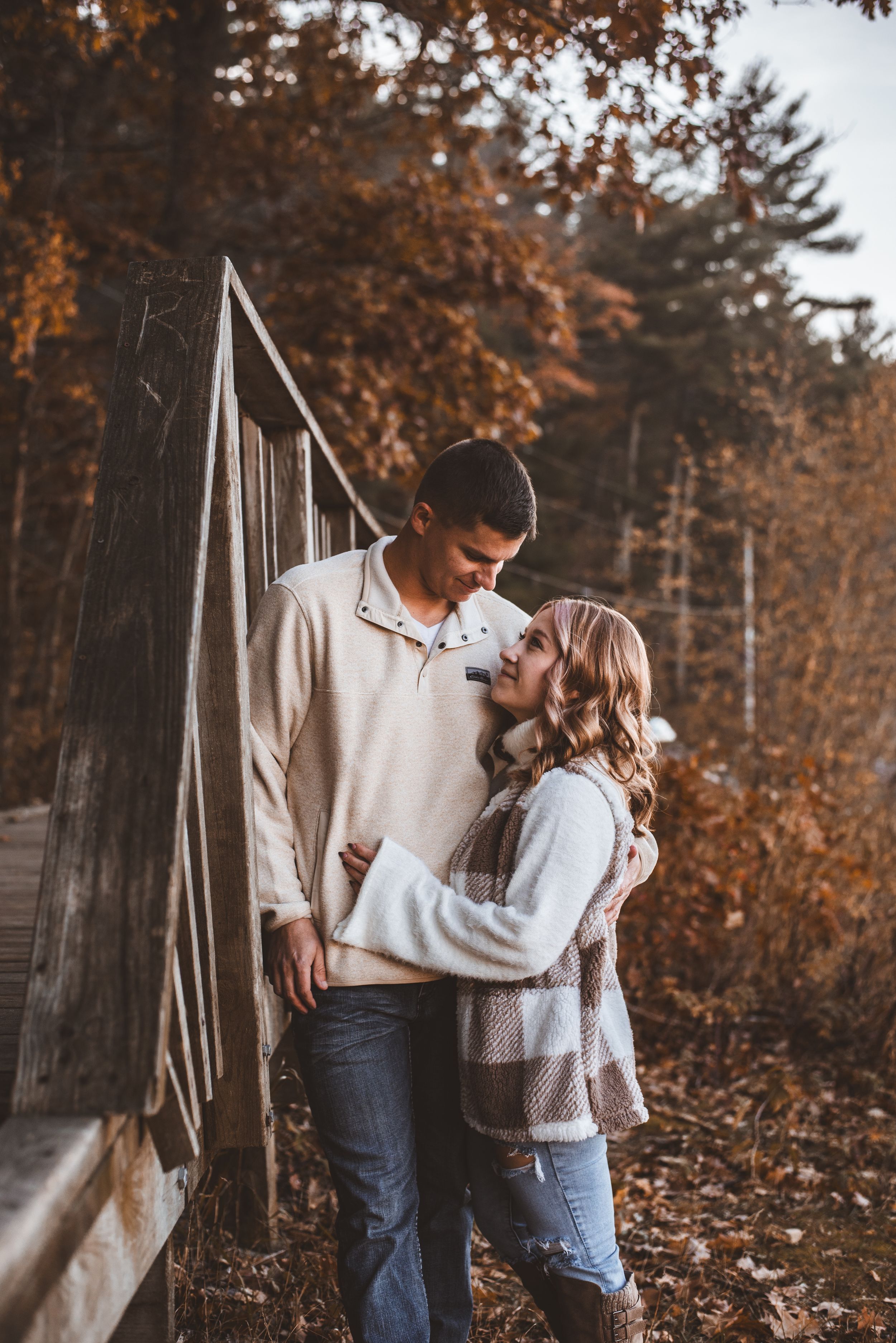 Couple sharing a tender moment against rustic bridge railing with fall foliage in background.