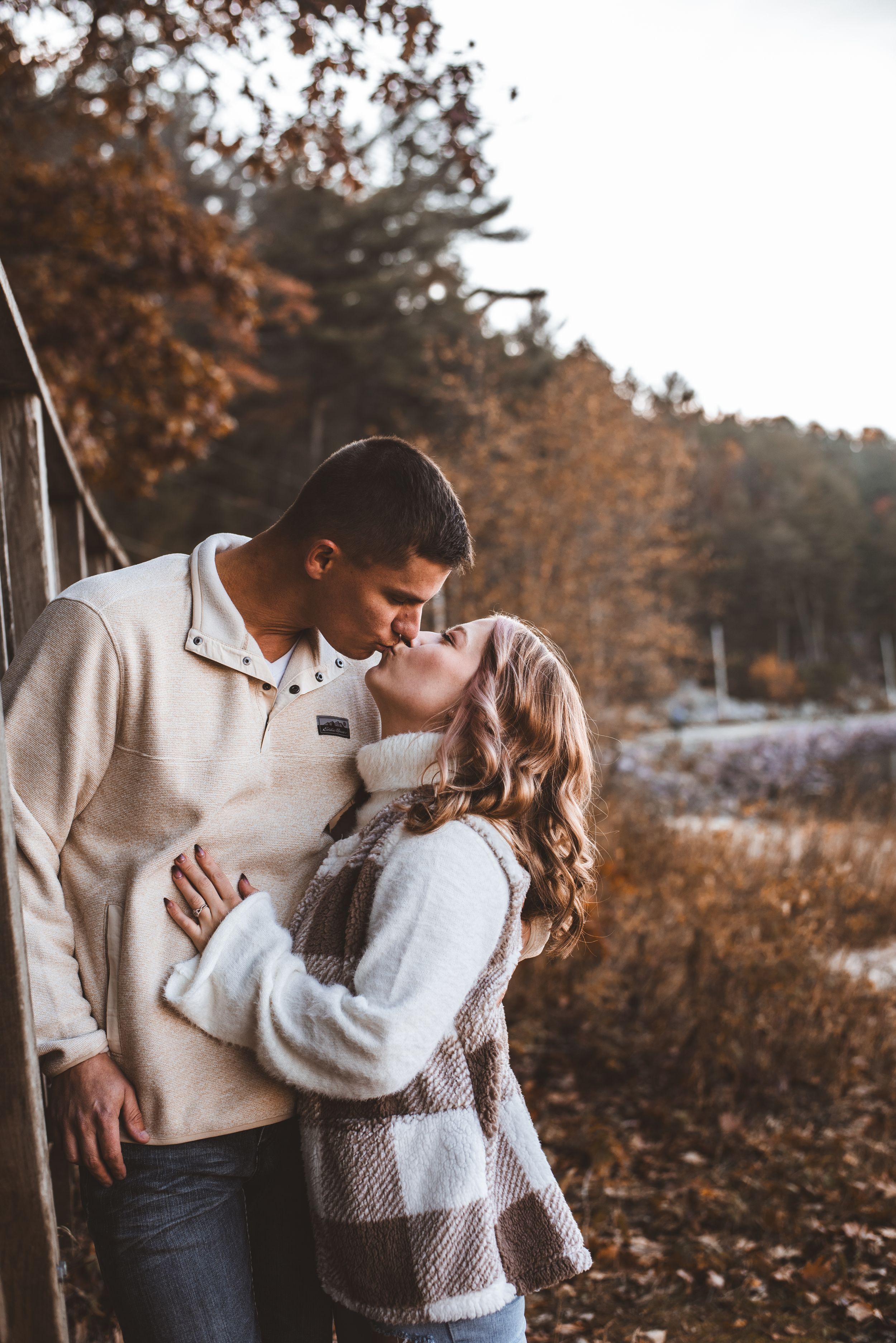 Intimate couple moment by wooden bridge railing surrounded by autumn trees and lakeside scenery.