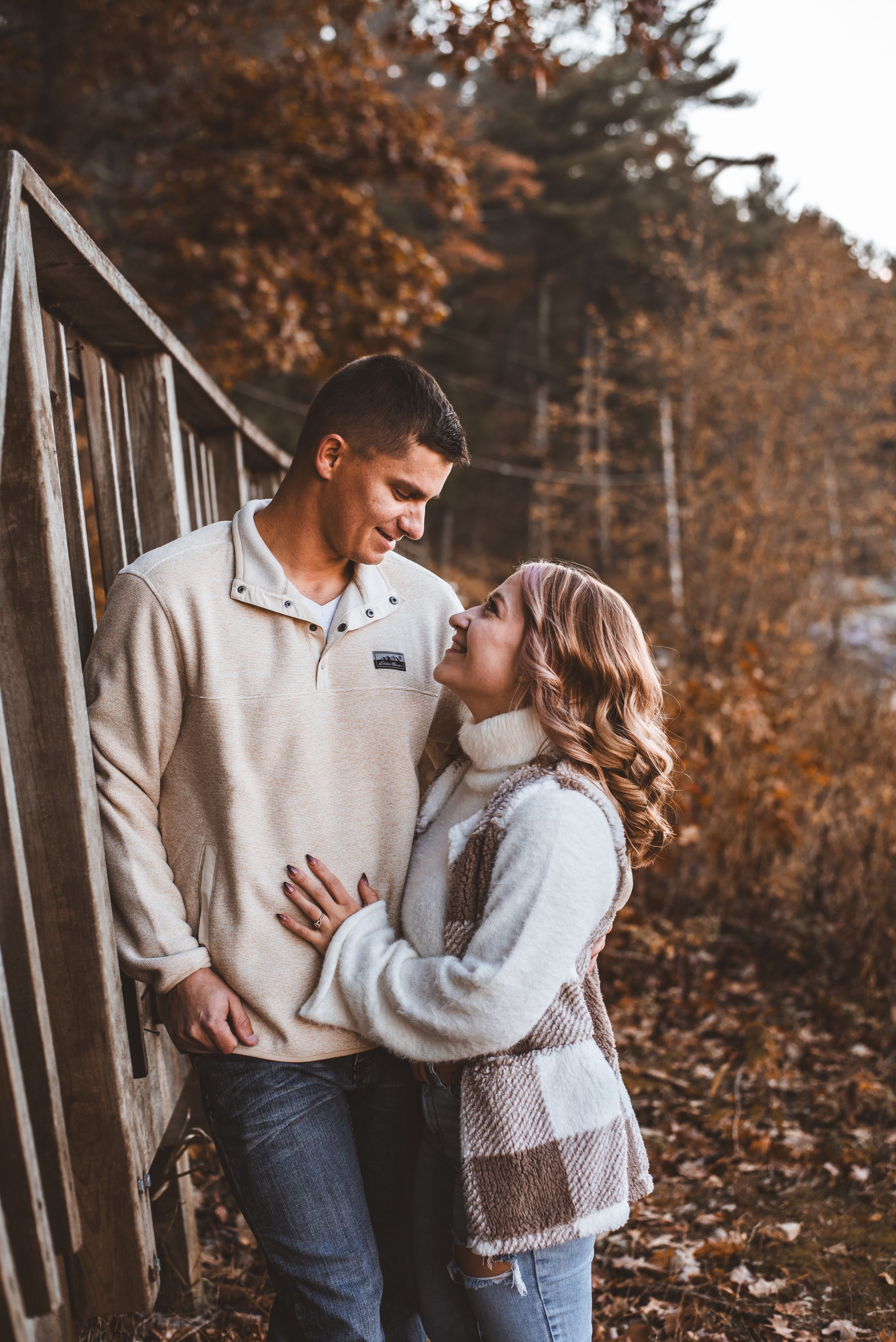 Couple gazing at each other by wooden railing with fall colors and lake in background.