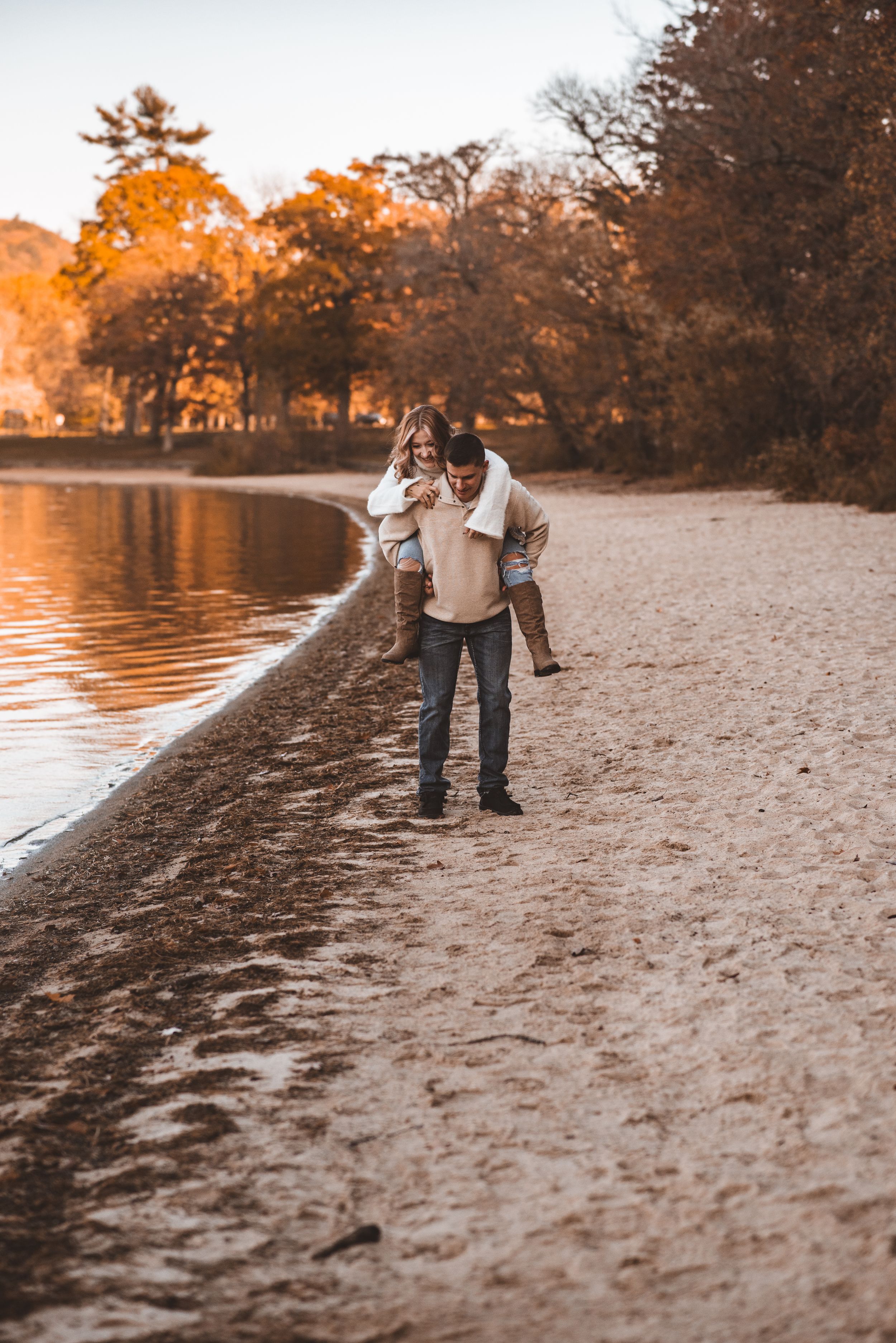 Couple playing on beach at golden hour with autumn trees and lake reflecting sunset light.