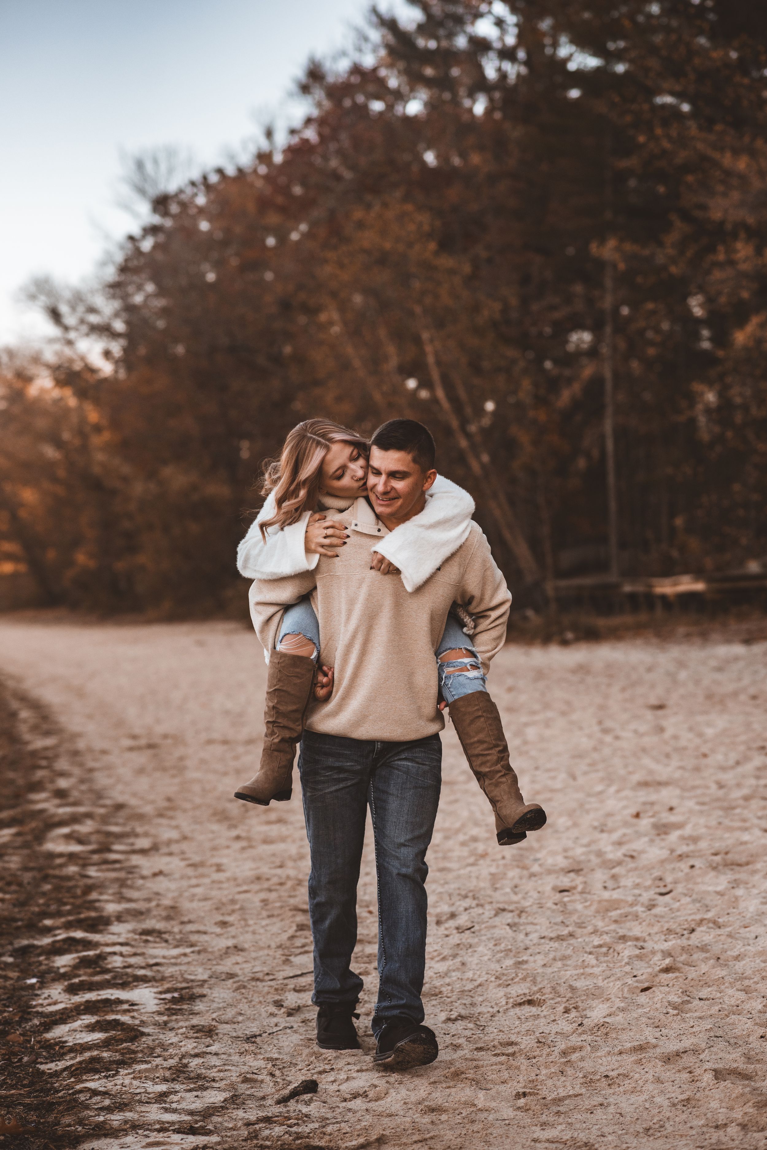Couple enjoying playful piggyback ride on beach path with fall foliage and lake in background.