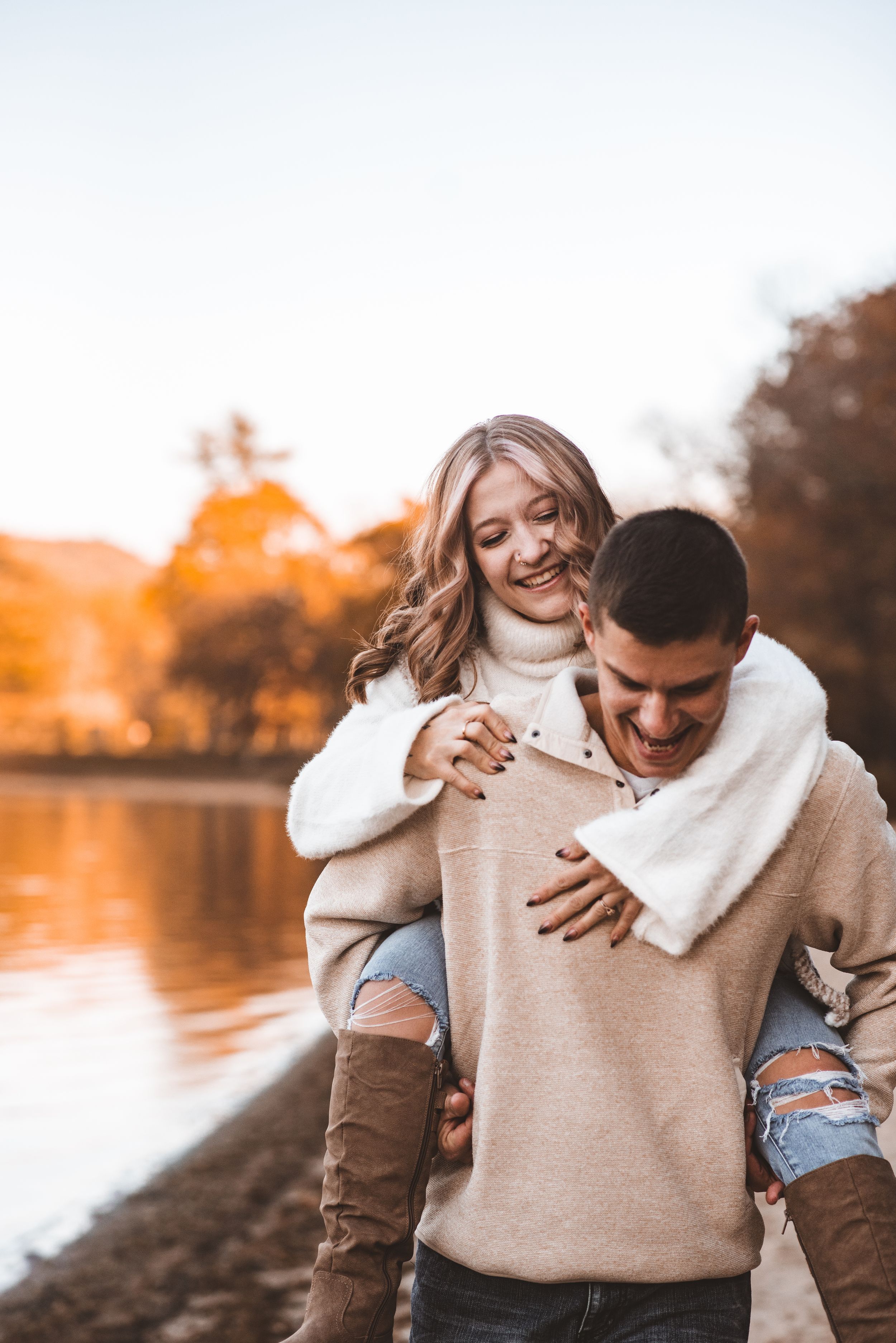 Couple sharing joyful laughter during piggyback ride by lake at sunset.