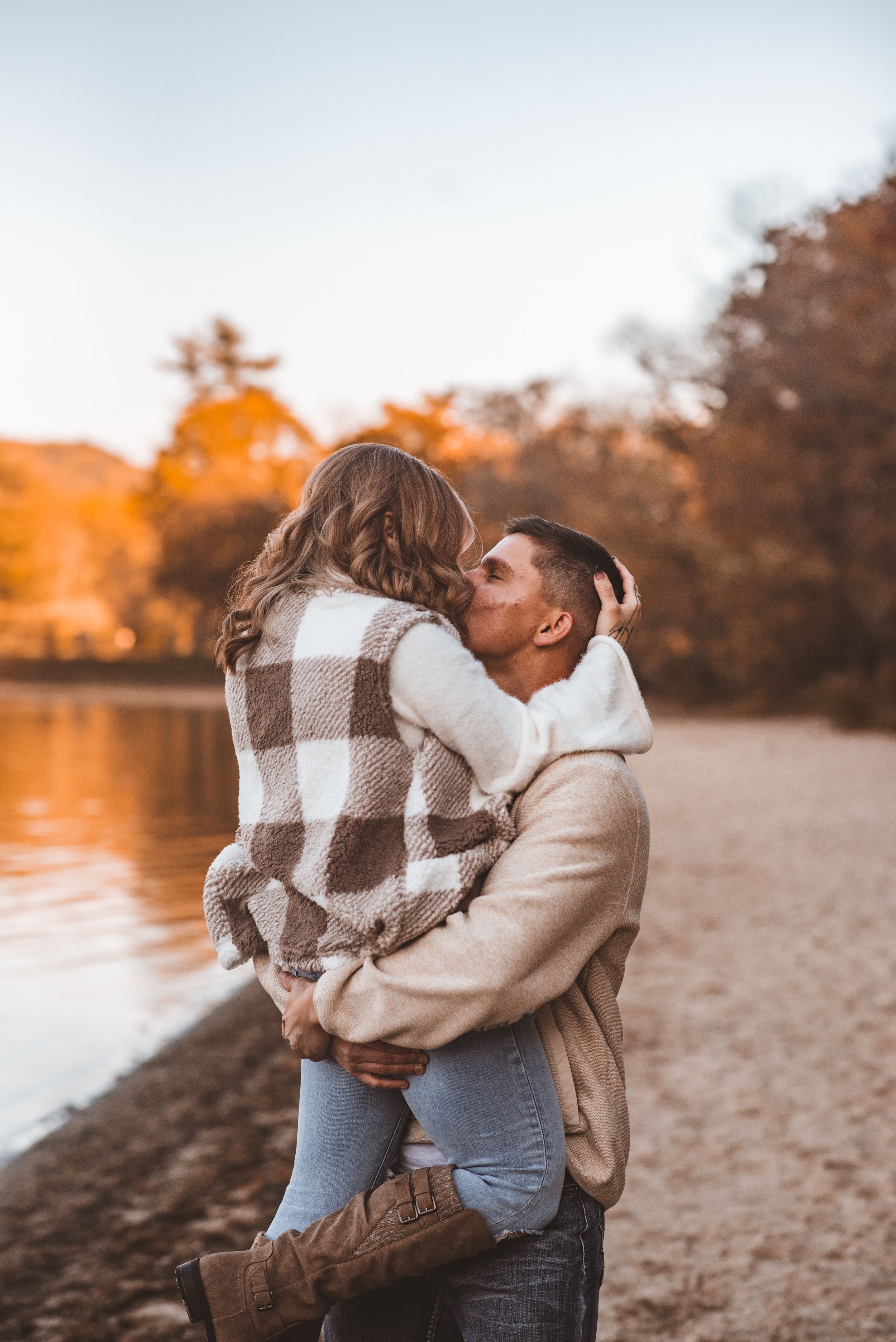 Romantic embrace between couple on sandy beach with autumn trees in background.