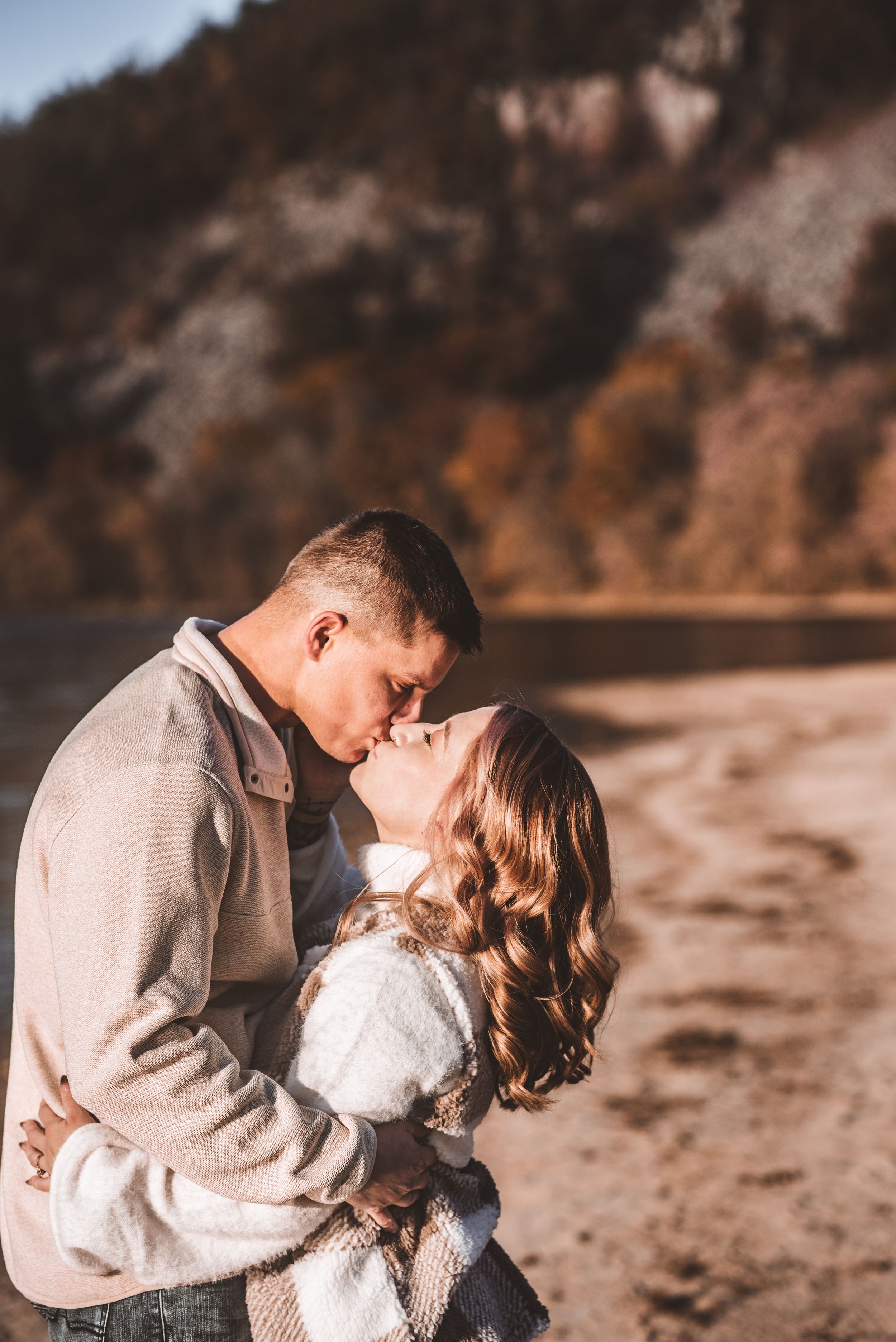 Close embrace between couple on beach with autumn foliage and lake in background.