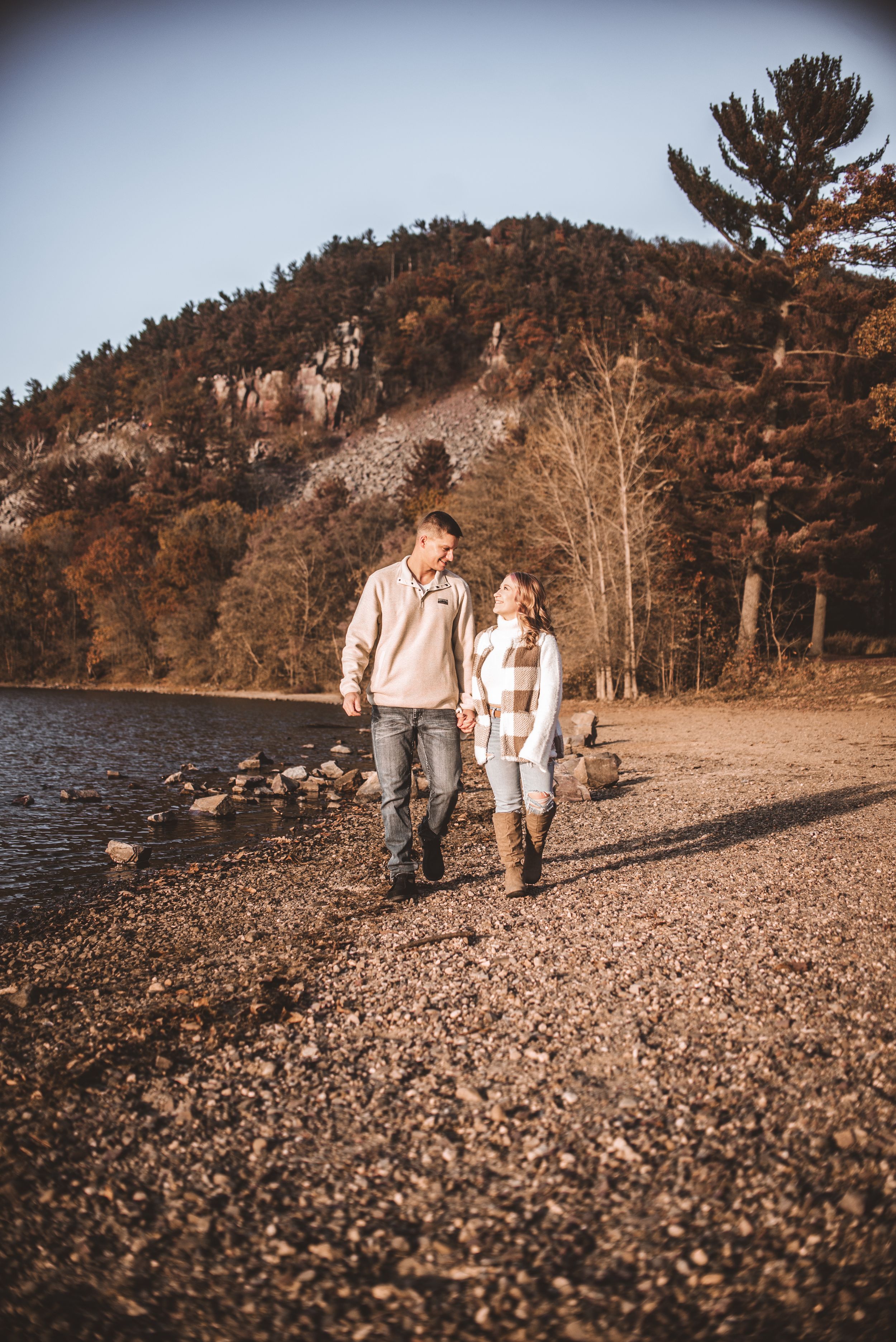 Walking hand in hand along pebbly lake shore with autumn hills in background.