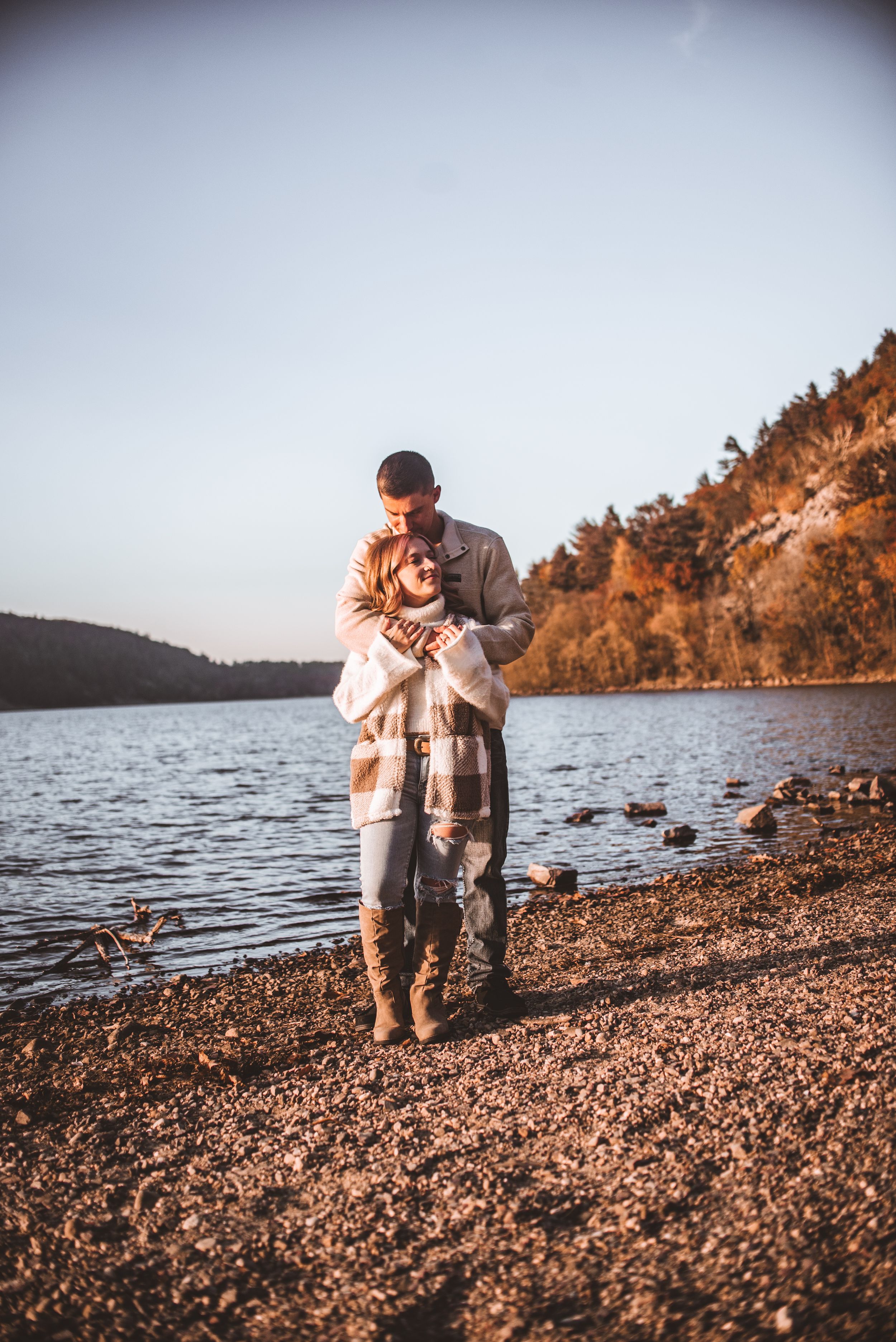Warm embrace on rocky beach with lake and autumn foliage in background.