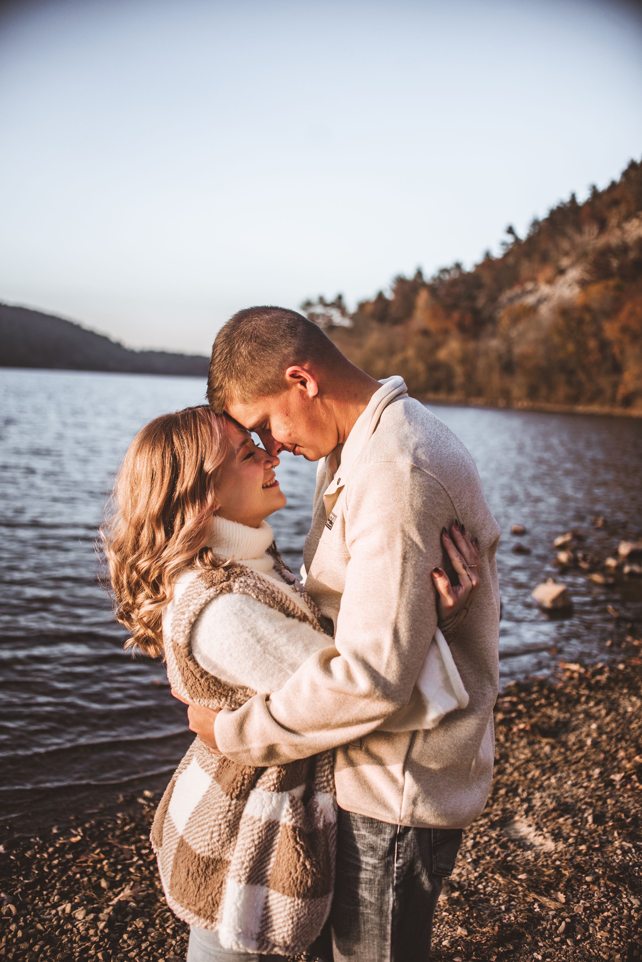 A tender lakeside moment captured during golden hour with warm neutral tones and rocky shoreline.