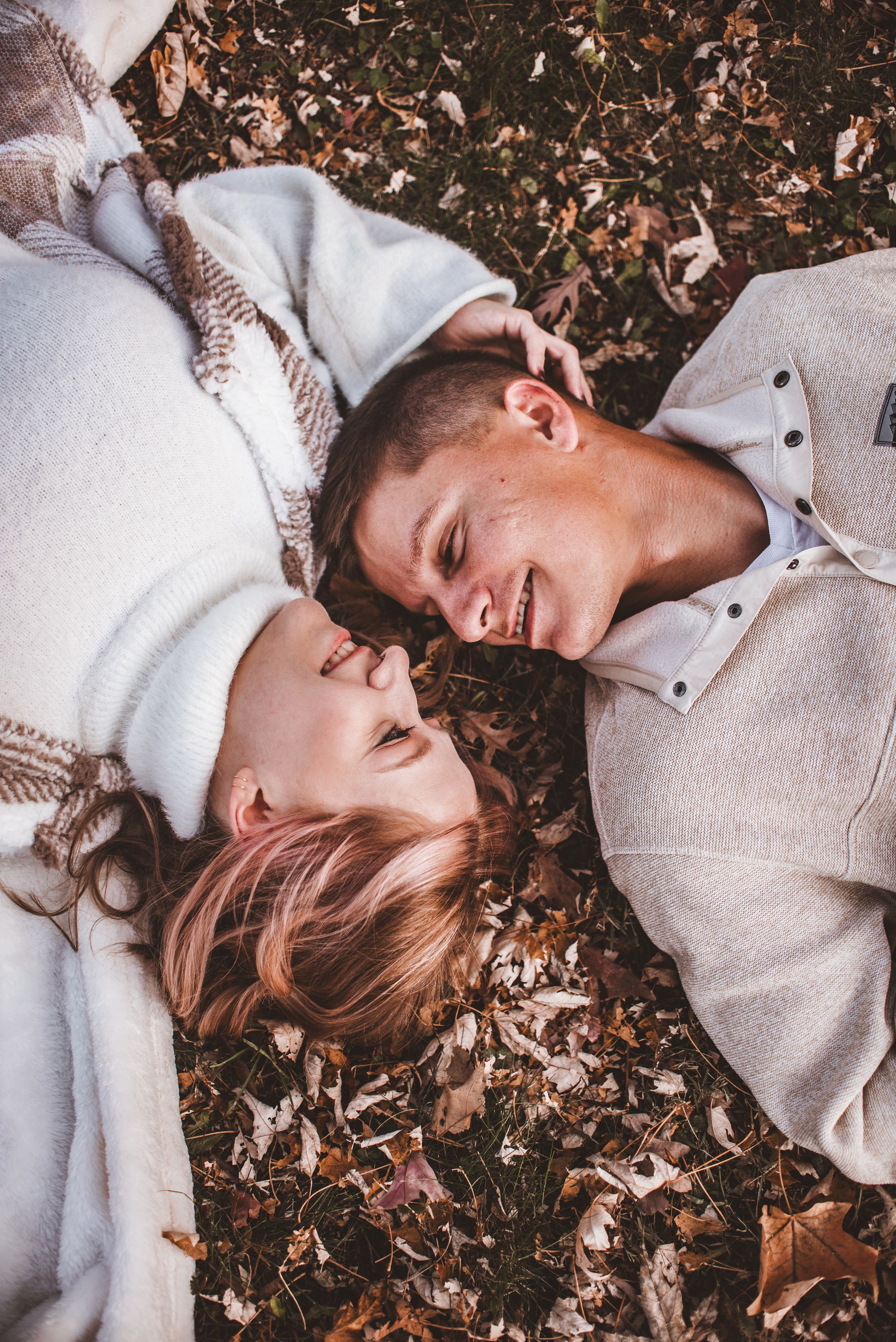 A joyful overhead portrait of a couple lying in autumn leaves wearing matching light-colored sweaters.