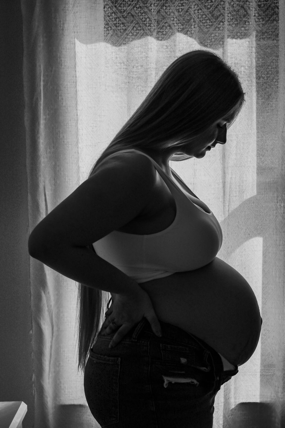 Black and white maternity portrait showcasing an expecting mother standing in profile beside a window.