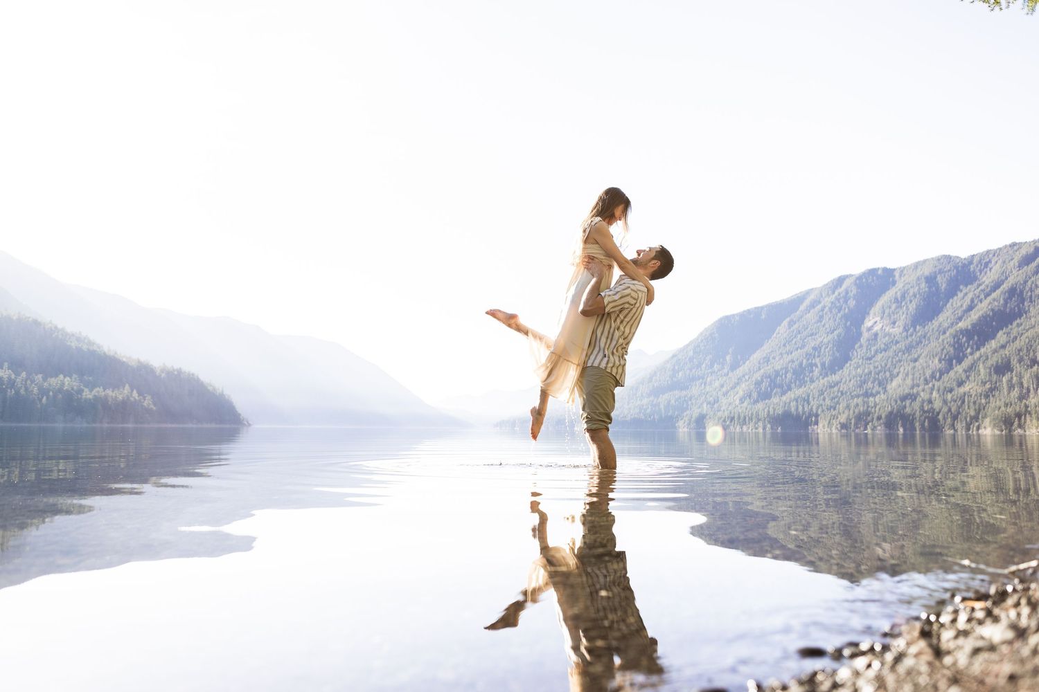 A person stands with outstretched arms on a rocky cliff overlooking a scenic mountain lake landscape.
