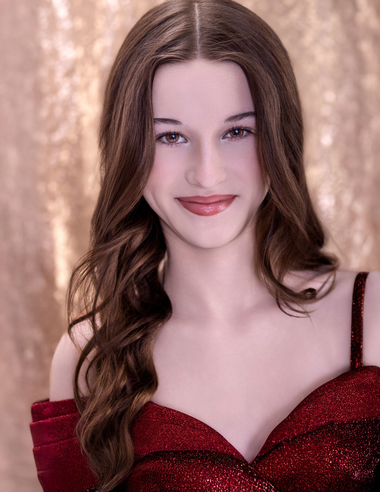 Portrait in a red off-shoulder dress against a sparkly gold backdrop with flowing brown wavy hair and natural makeup.