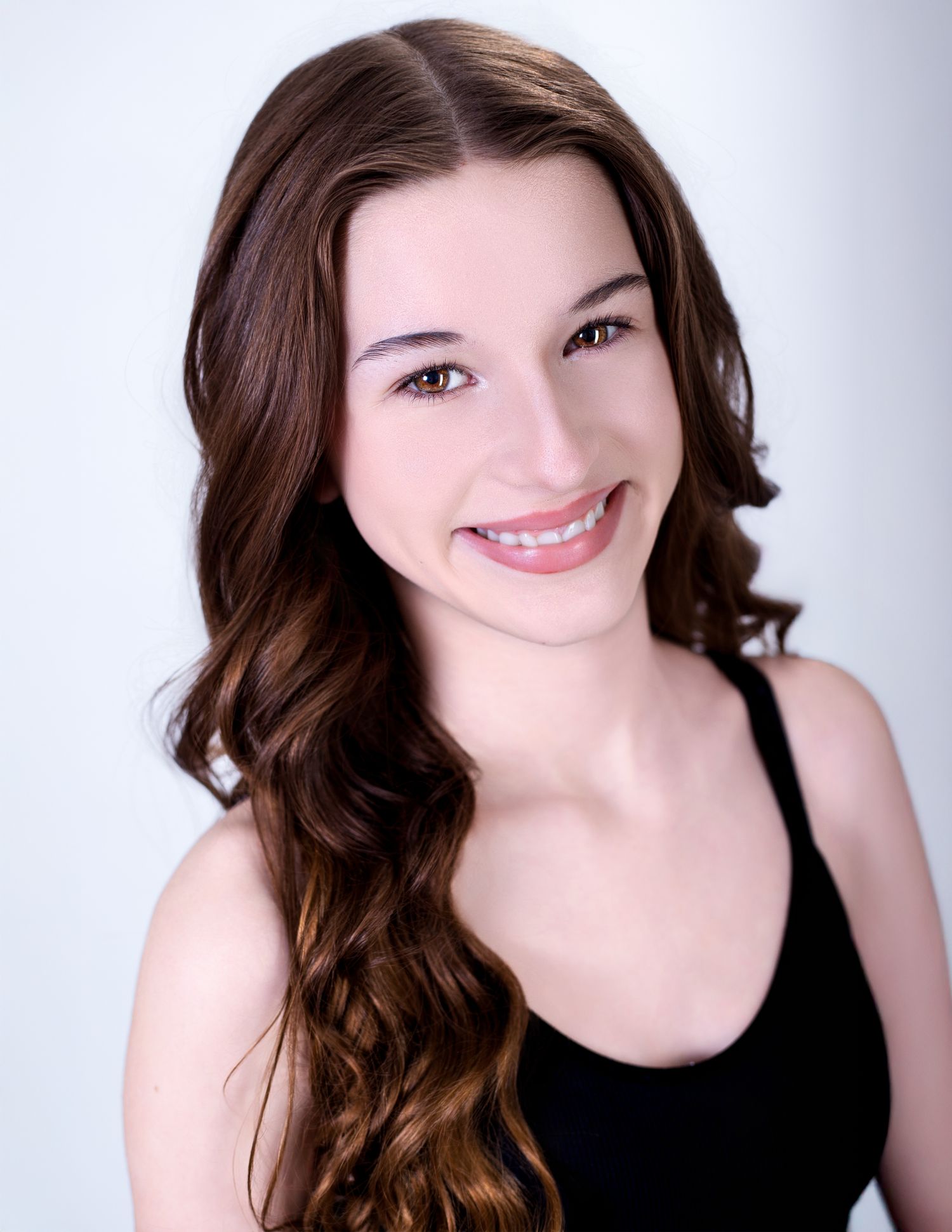 Professional headshot of a person with long brown wavy hair wearing a black tank top against a light gray background.