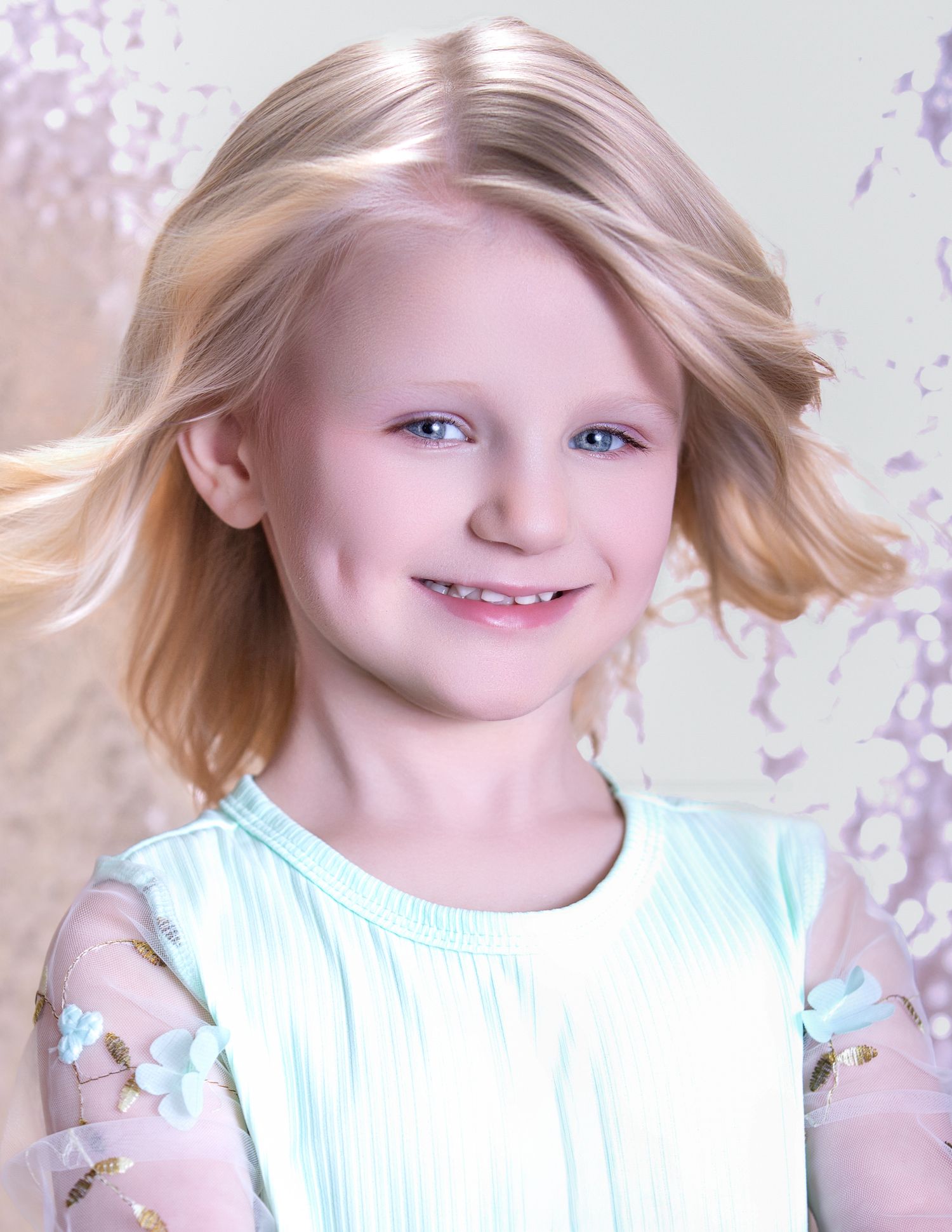 Smiling young blonde girl in a mint green floral dress with sheer sleeves, posed for a portrait photo.