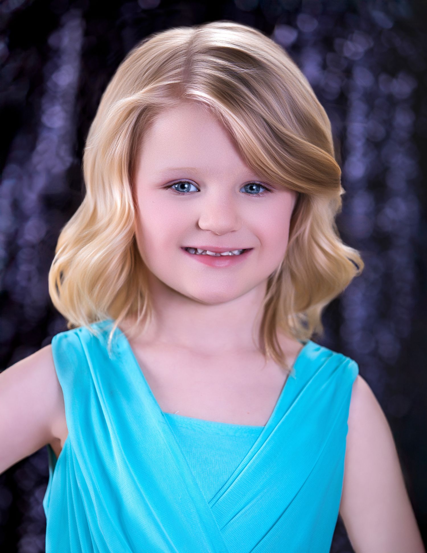 Young girl with blonde wavy hair and blue eyes smiling, wearing a turquoise dress against a dark backdrop.