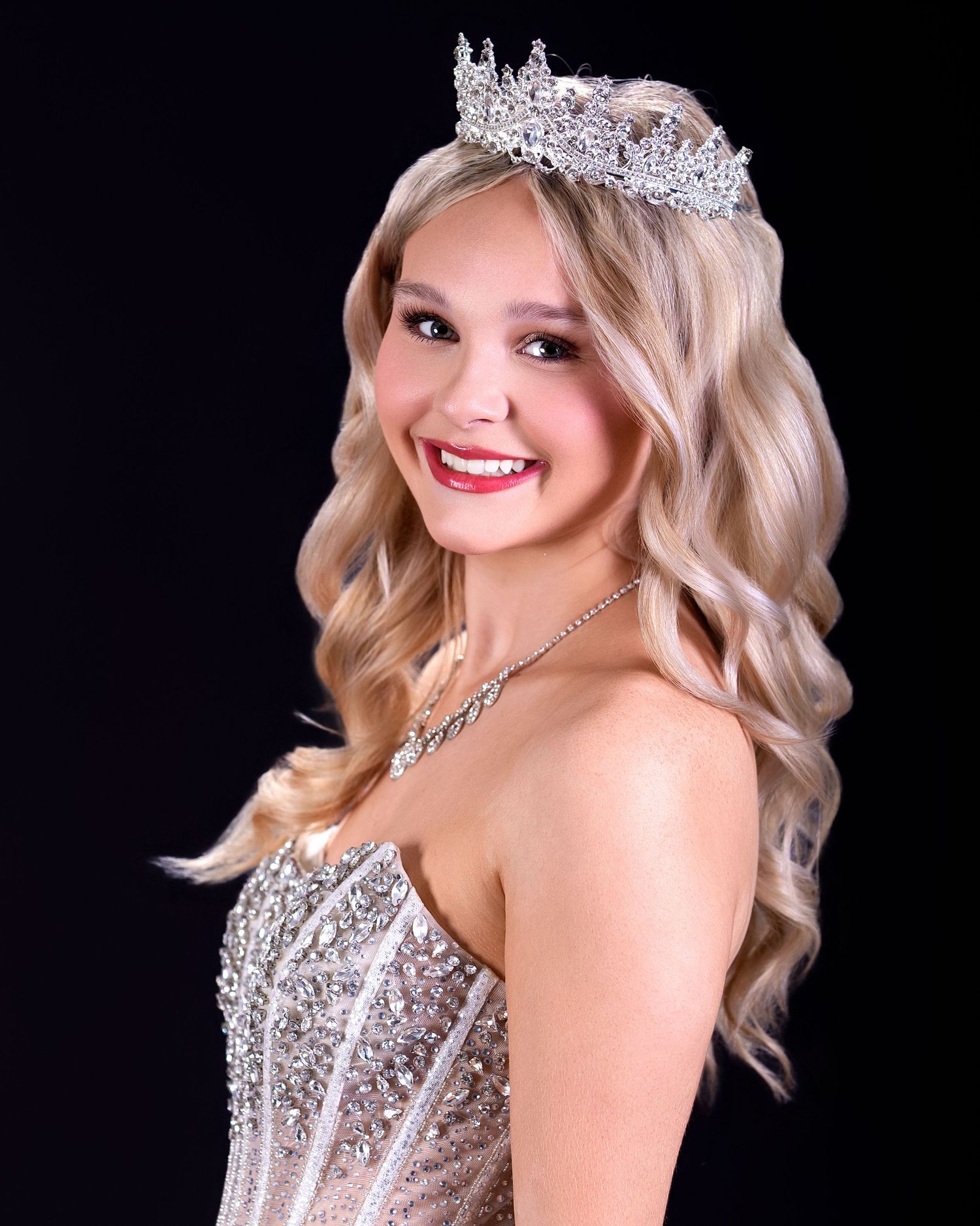 Young woman wearing a sparkling tiara and sequined gown, smiling confidently against a black background.