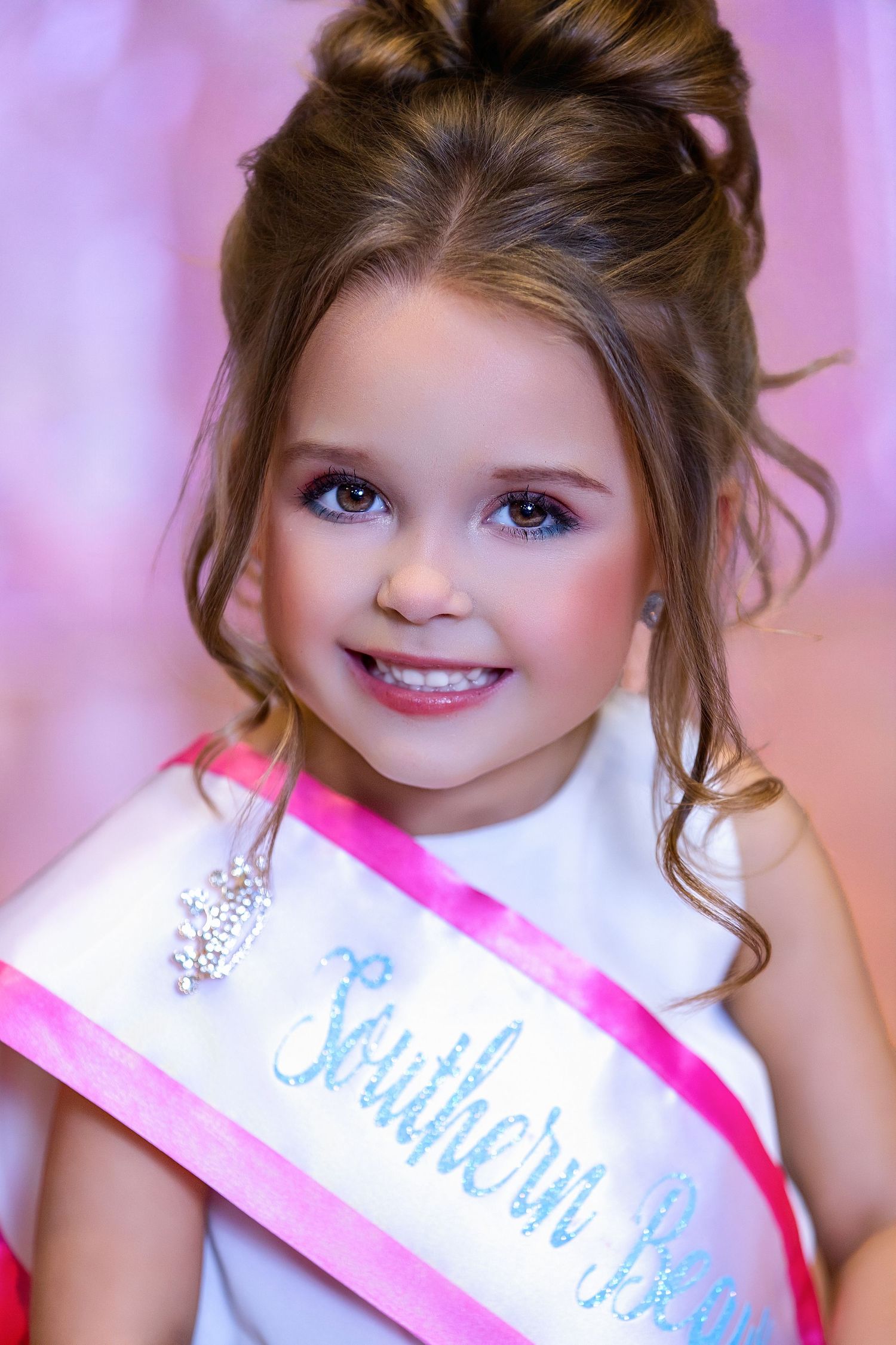 A young pageant contestant wearing a white dress and pink sash smiles brightly against a pink backdrop.