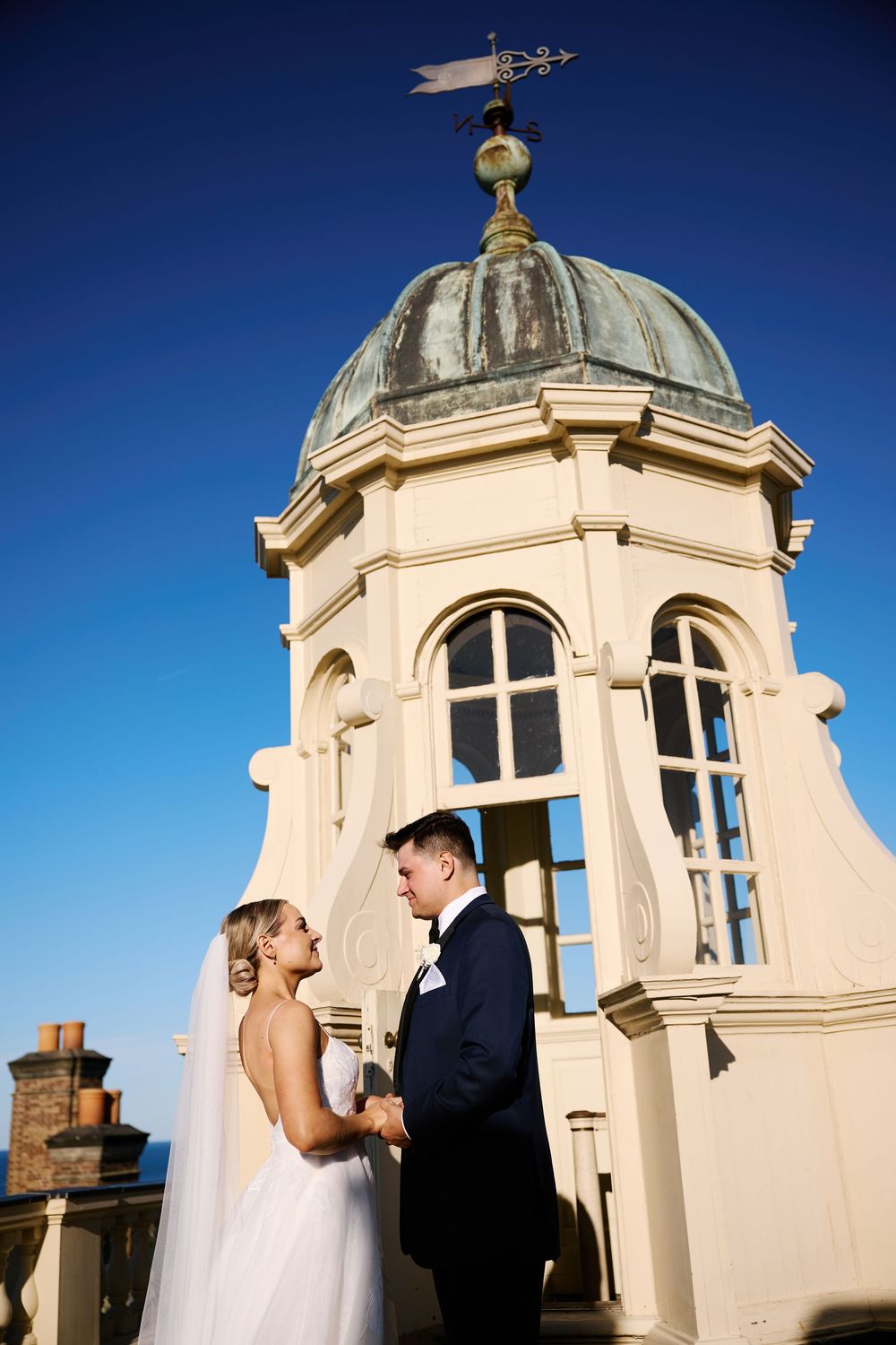 A wedding couple stands against a white domed architectural structure under bright blue sky in a romantic sequence.