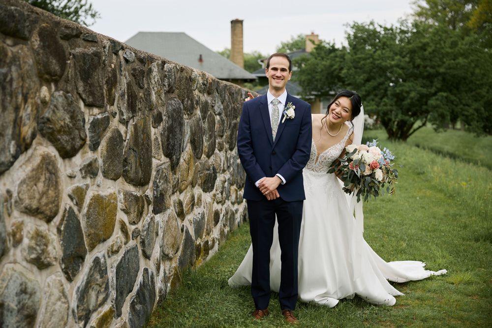 A couple poses for outdoor wedding photos in a rustic natural setting with stone walls and greenery.