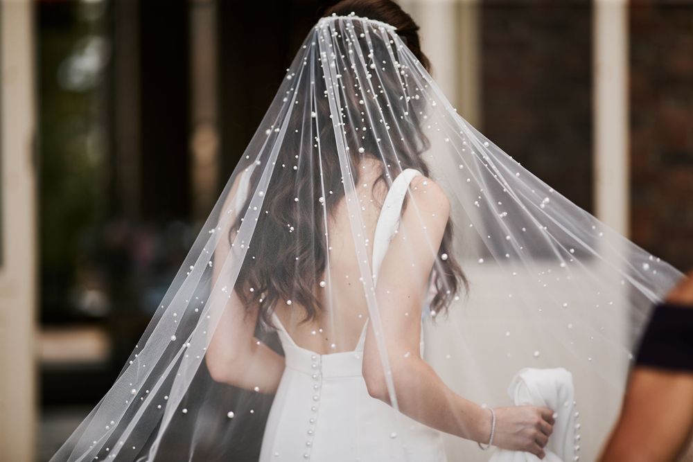 Bride in a flowing white veil and gown poses against a wooden wall with natural lighting.