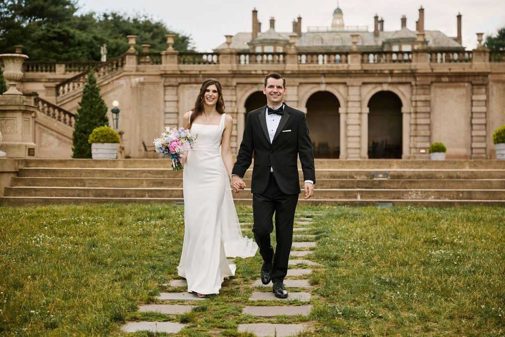 A couple in wedding attire walks together on the grass outside a grand historic stone building.