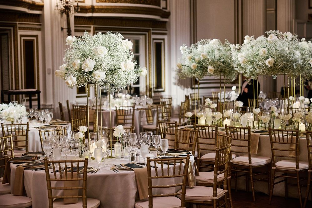 Elegant wedding reception hall with white floral centerpieces and gold chiavari chairs around circular tables.