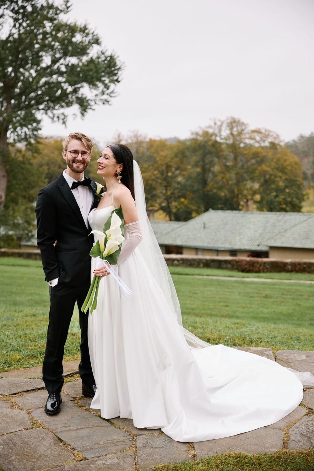 A bride and groom share a joyful moment together outdoors on their wedding day on a stone path surrounded by autumn trees.