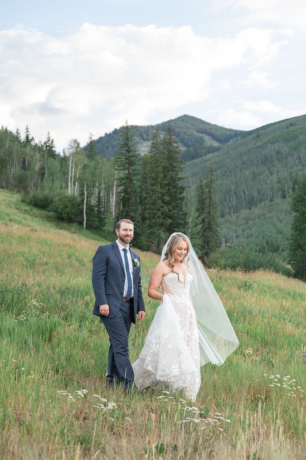 Bride and groom walking through a mountain meadow, bride in lace gown with flowing veil, groom in navy suit.