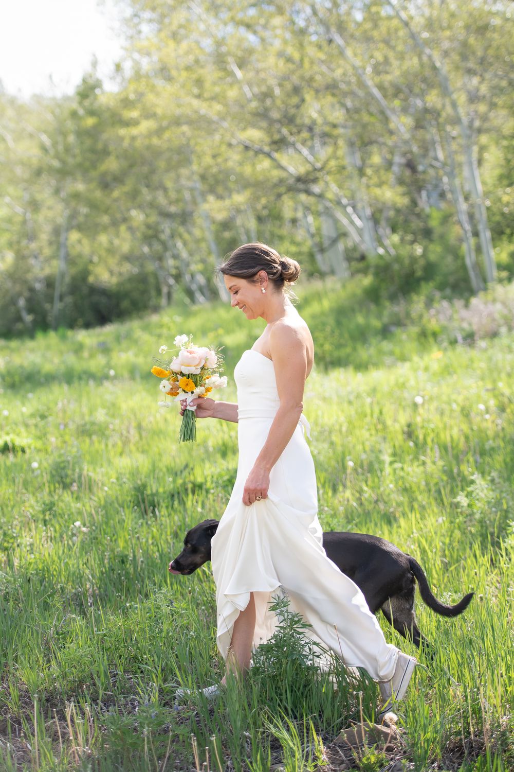 Bride in white gown holds colorful bouquet while walking with black dog through lush green meadow.