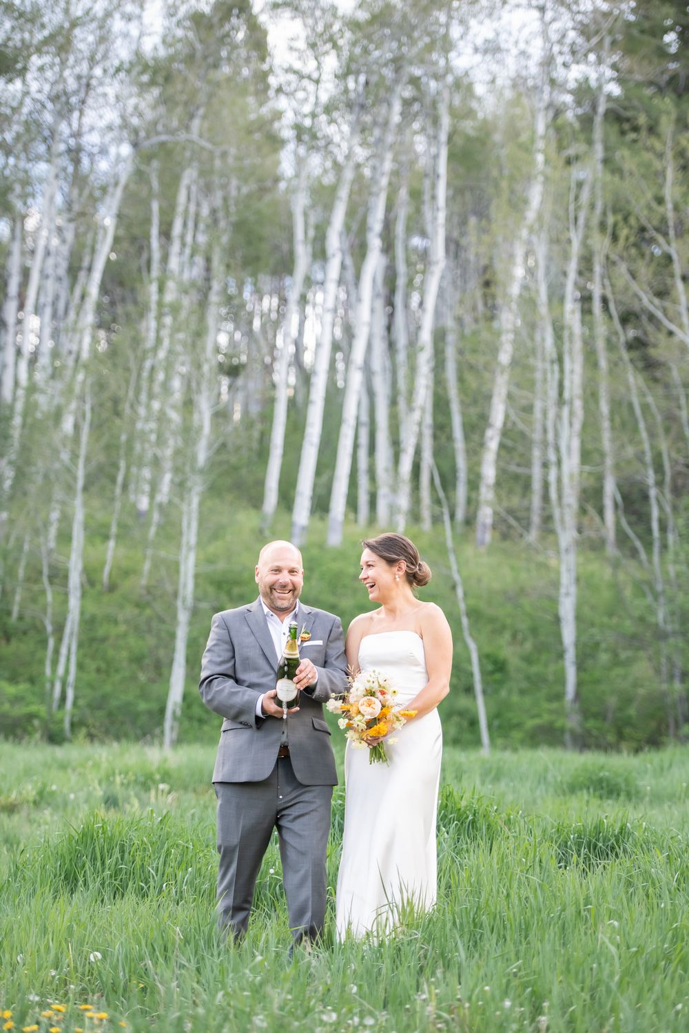 Bride and groom laughing in a green meadow with white birch trees, holding champagne and a yellow bouquet.