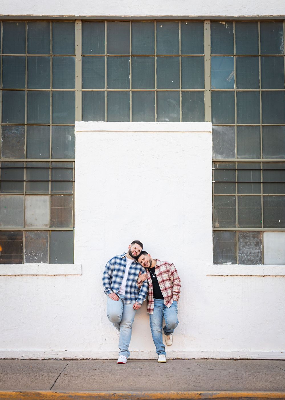 Two people in plaid shirts lean against a white brick wall with industrial windows outdoors.