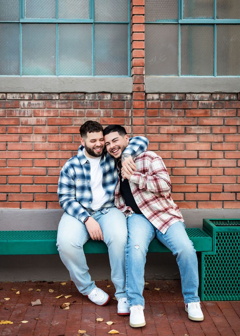 Two men in plaid shirts laughing together on a green bench against a brick wall outdoors.