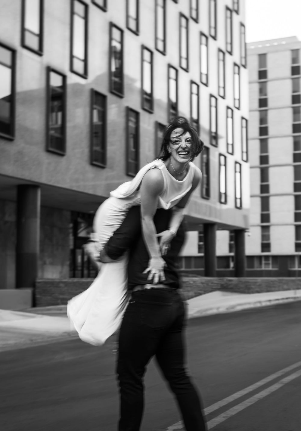 Black and white photo of a laughing woman being carried by a man on a city street near modern buildings.