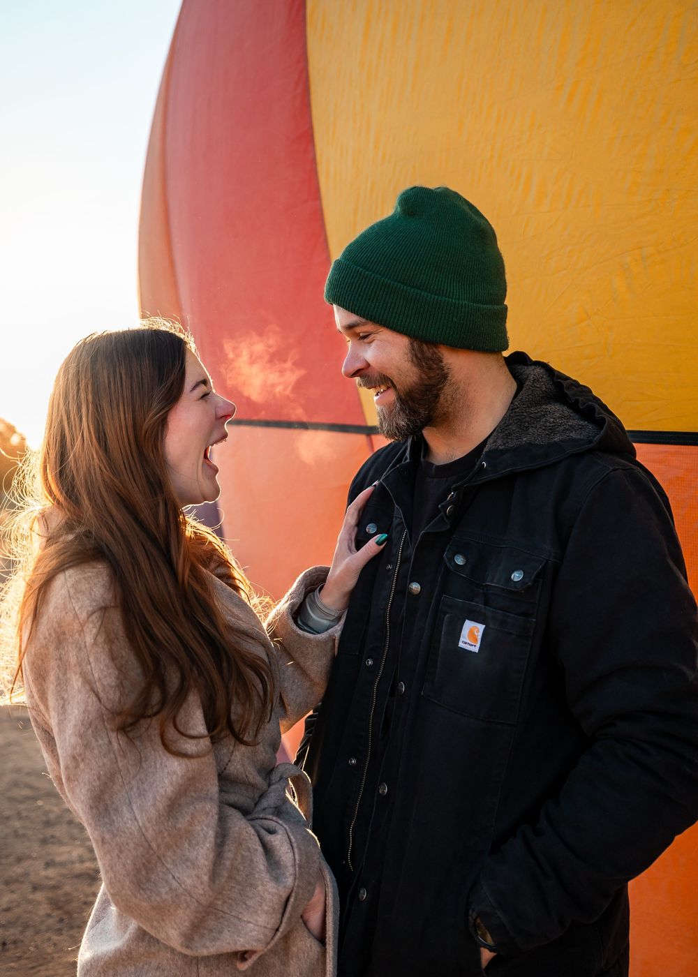 A couple in winter attire shares a tender moment against a vibrant orange and pink wall at sunset.