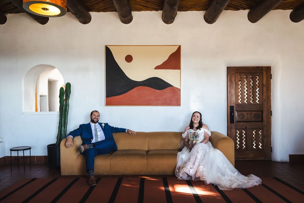 A series of people seated in rustic wooden chairs line up against an adobe-style wall with painted artwork and decor.