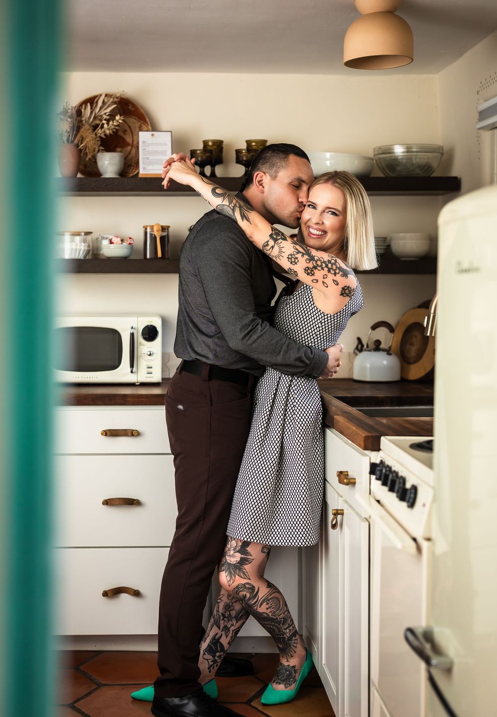 A couple shares an intimate moment together in a cozy white kitchen while standing near the refrigerator.