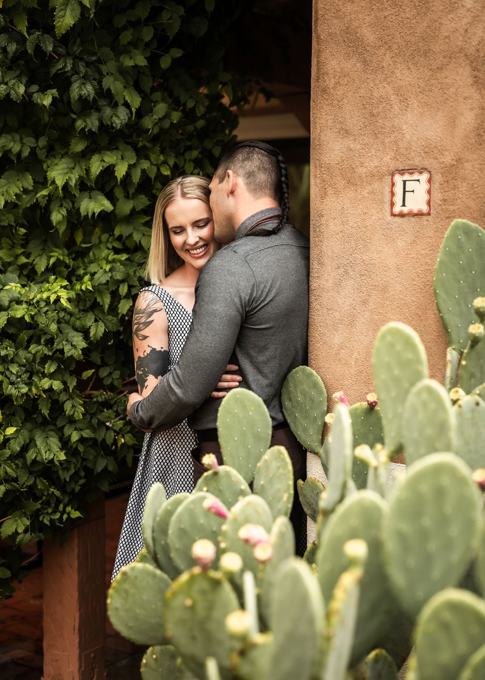 A romantic couple embraces near an adobe wall with green cactus plants and ivy in the foreground. Albuquerque, New Mexico