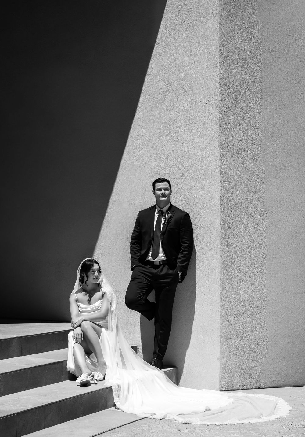 Black and white wedding photo of bride sitting on steps and groom leaning against wall in dramatic light.