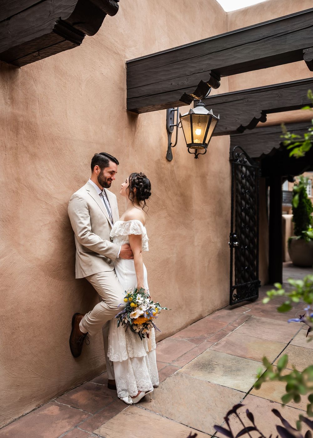 A couple in white wedding attire shares an intimate moment against an adobe wall in Santa Fe style architecture.