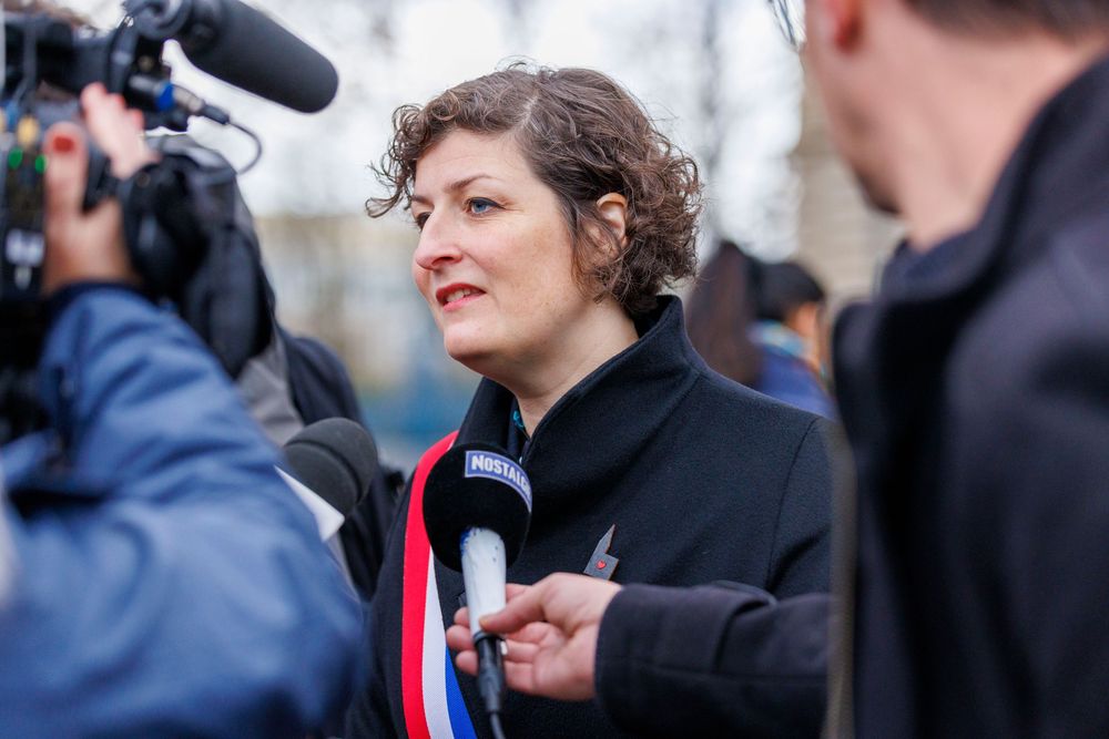 Someone wearing a French tricolor sash speaks to media with microphones and cameras at an outdoor press event.