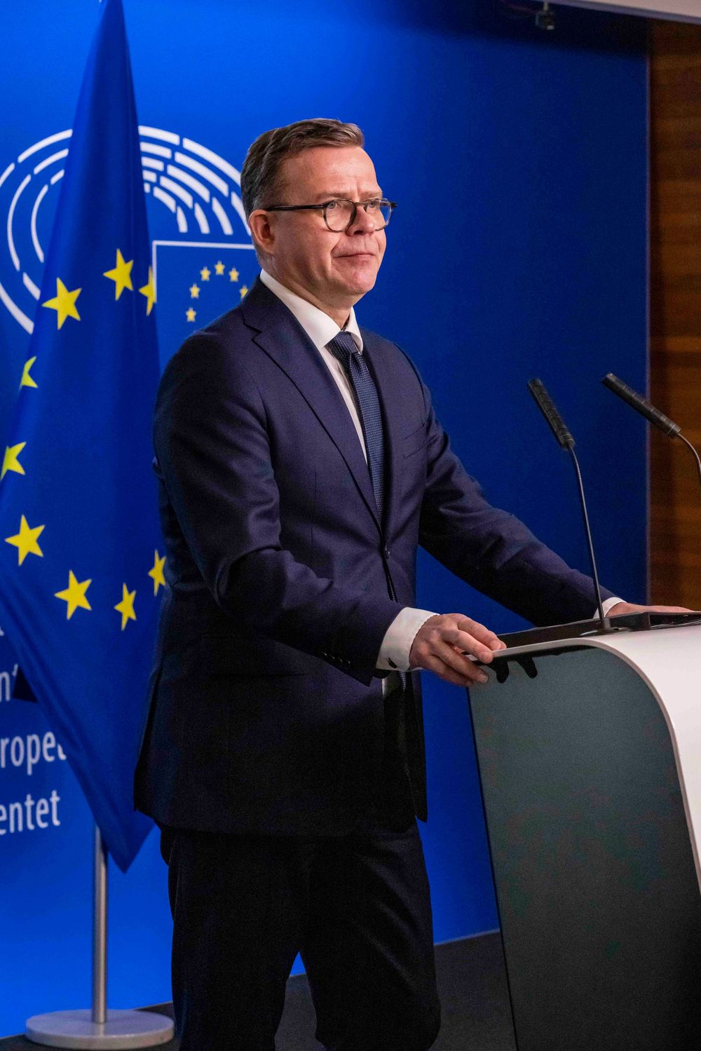 European Commission official speaking at a press conference podium with EU flags in the background.